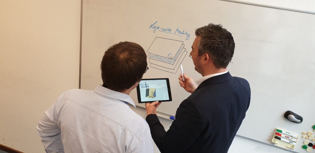 Picture of two men in front of a whiteboard discussing a part that is shown on a tablet one of them is holding